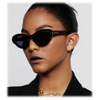 Woman wearing black cat-eye sunglasses and gold jewelry on a white background
