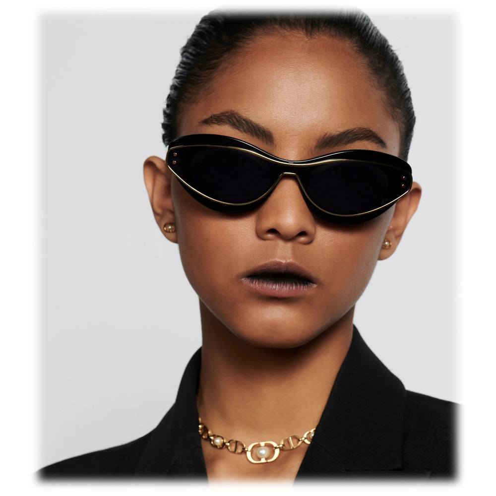 Woman wearing black cat-eye sunglasses with gold trim, gold chain necklace, and black blazer